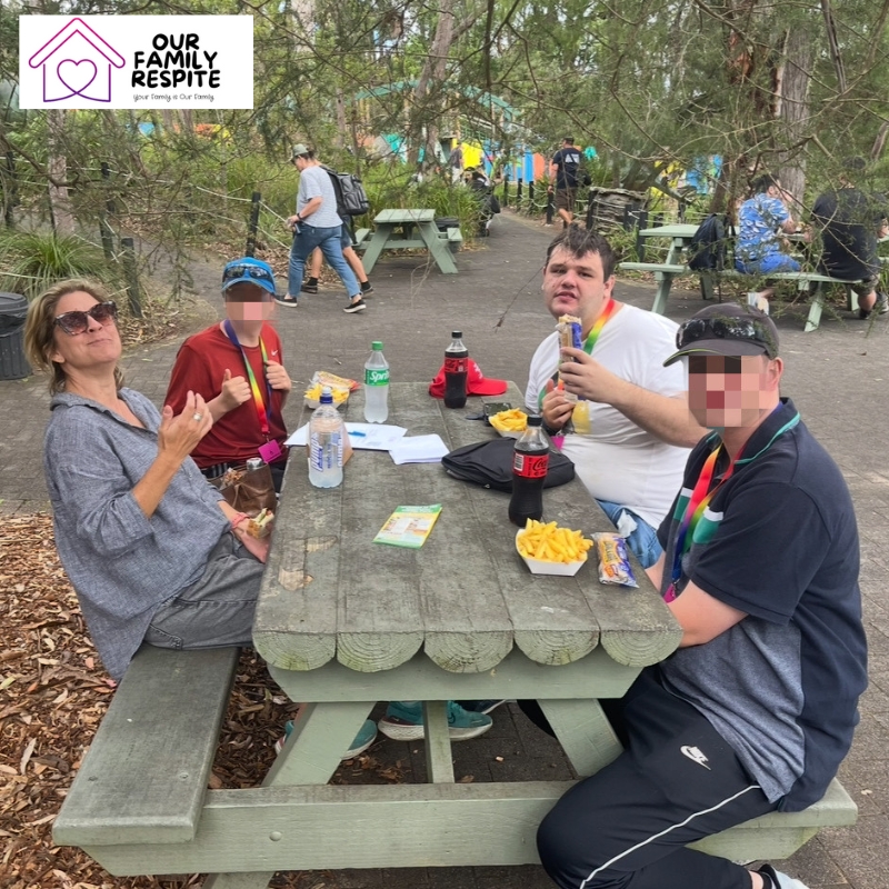 Families enjoying school holiday care for children with disabilities at a picnic table in a vibrant park, sharing food and drinks together.