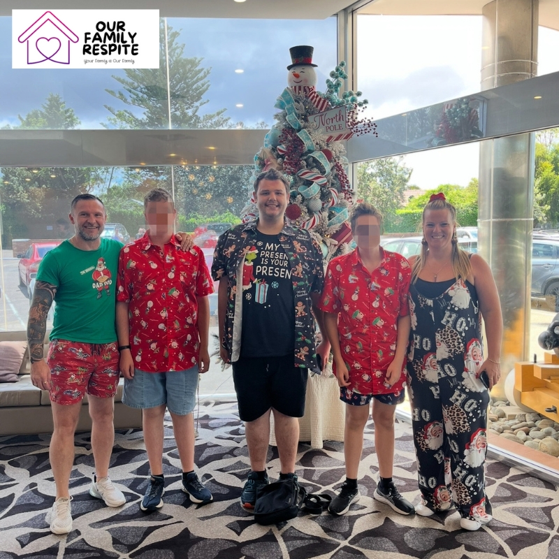 A cheerful moment from School Holiday Respite Care with five people in holiday outfits by a colourful Christmas tree.