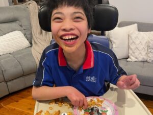 Smiling boy in support chair enjoying food during respite programs for youth in a home setting with familiar surroundings