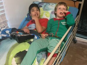 Two boys in pyjamas lying in bed with a communication device, representing down syndrome respite care in a home setting.