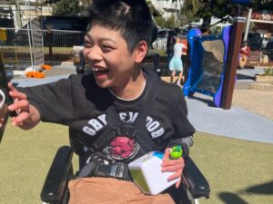 Smiling boy in wheelchair holding a toy at an outdoor playground, enjoying flexible respite care with support nearby