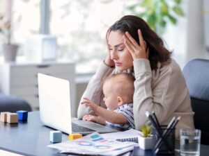 A stressed mother works on her laptop with her baby on her lap, highlighting need for a respite care program.