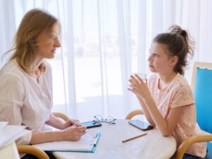 A female counsellor talks with a young girl during a respite care program session, clipboard and notes on table.