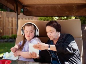 An older woman shows a tablet to a young girl with Down syndrome wearing headphones while sitting outdoors, illustrating respite care at home.